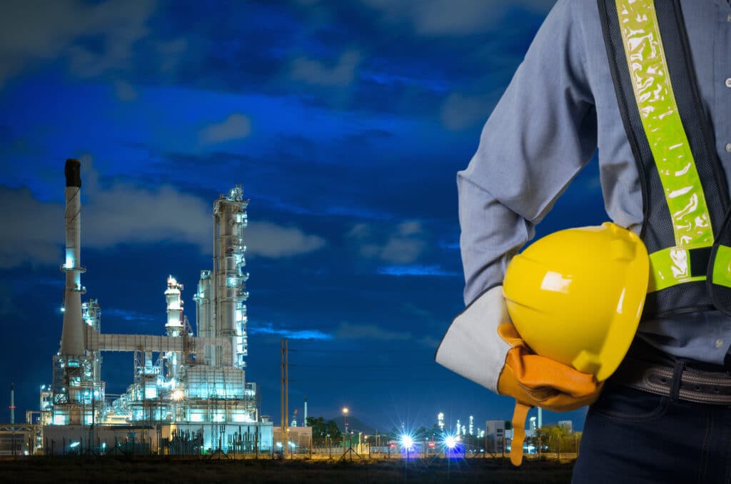 A person wearing high visibility safety gear and holding a hard hat stands in front of a refinery